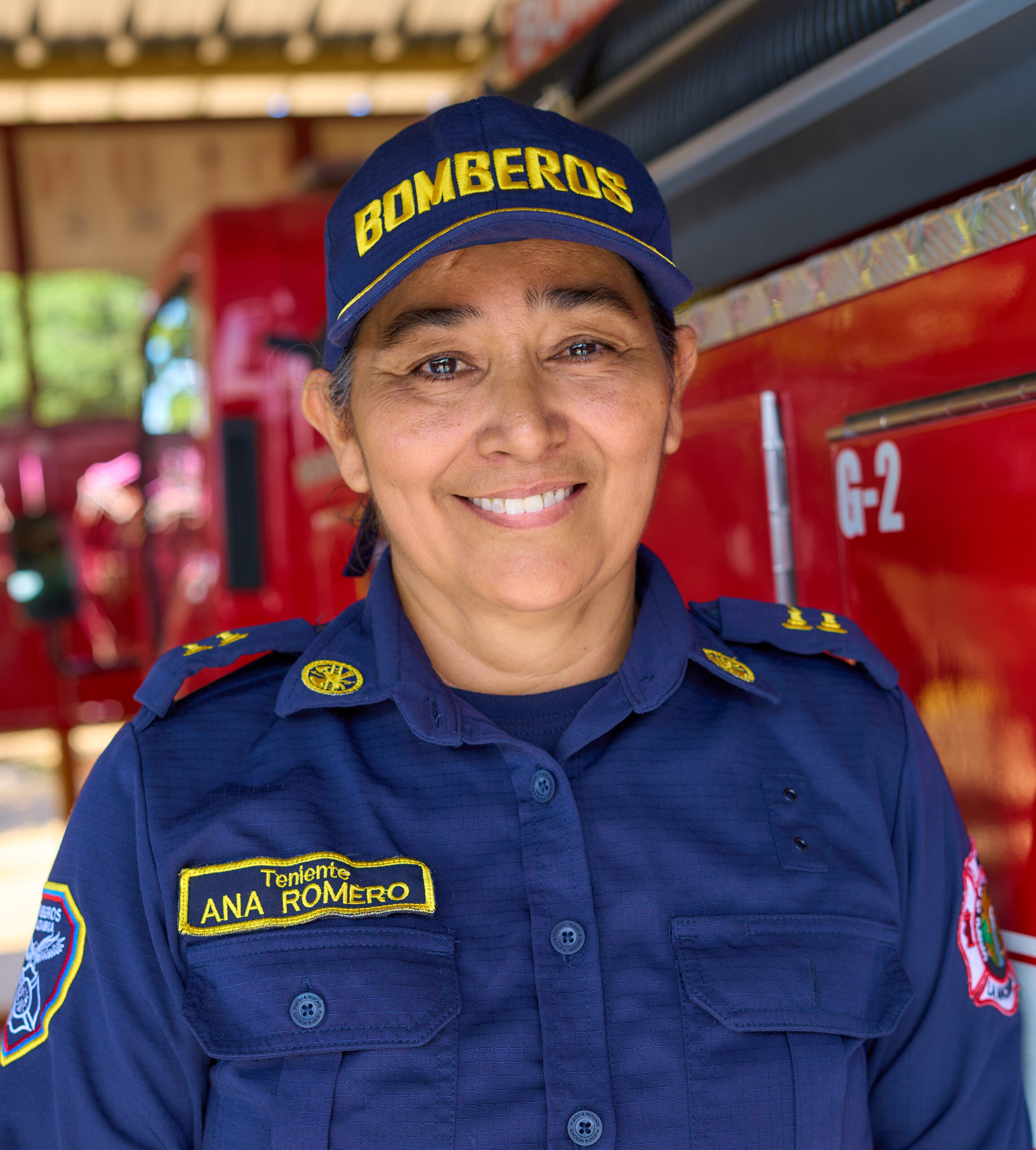 A Colombian female firefighter in blue uniform stands next to a red fire truck and smiles at the camera